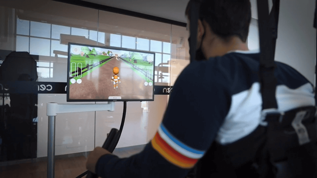 Una persona que lleva un arnés interactúa con un dispositivo de rehabilitación mientras juega a un juego que se muestra en un monitor en un entorno interior moderno.