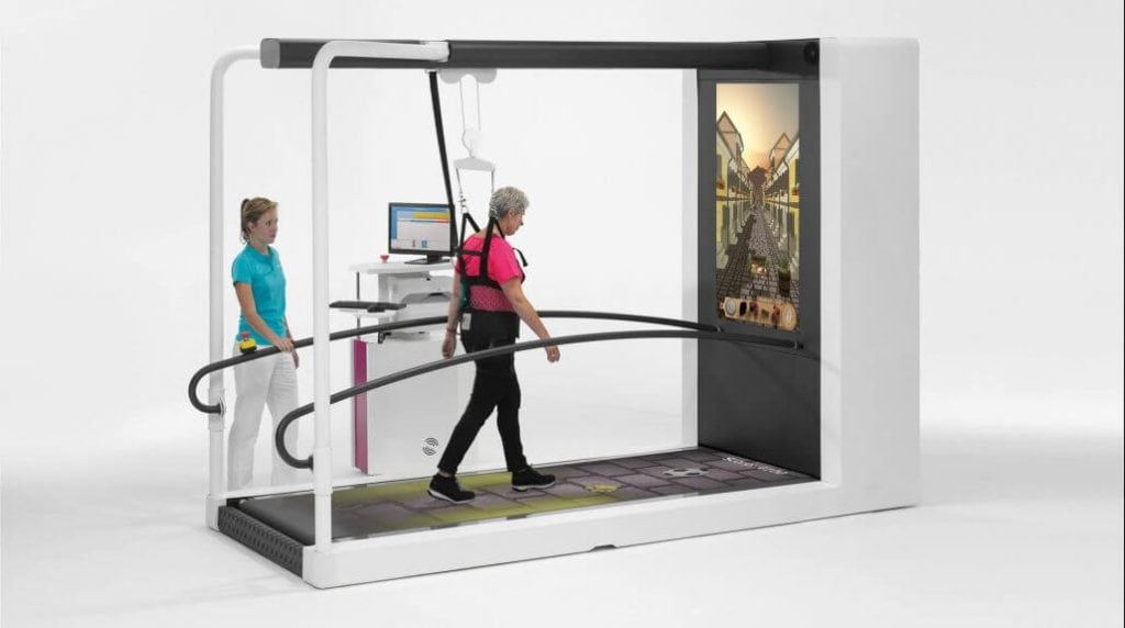 Un adulto mayor con un arnés camina sobre una cinta de correr con una pantalla de realidad virtual, supervisado por un clínico en un puesto informático cercano.