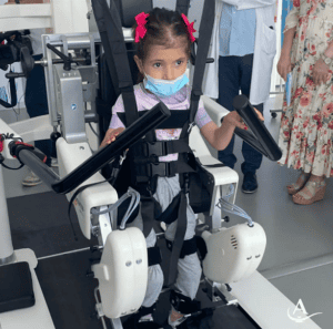 A young girl with a mask and pink hair bows uses a robotic exoskeleton device for physical therapy. Adults stand nearby, observing the session.