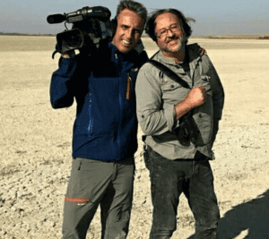 Two men stand on a dry, open landscape; one holds a video camera on his shoulder while both smile at the camera.