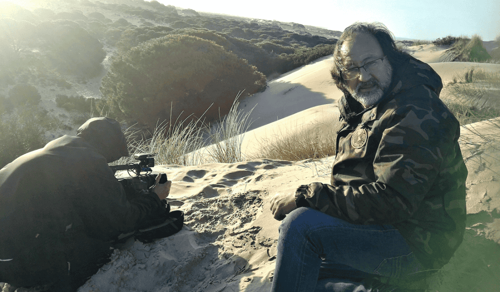 Dos personas en una duna de arena; una está filmando con una cámara mientras la otra está sentada frente a la cámara, con chaqueta y gafas. En el fondo se ven arbustos y dunas.