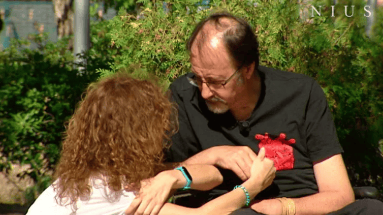 Una mujer de pelo rizado sujeta del brazo a un hombre sentado con gafas y camisa negra al aire libre, ambos parecen enfrascados en una conversación.
