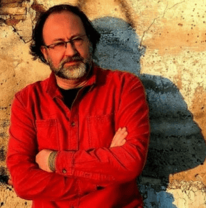 A man with glasses and a beard, wearing a red shirt, stands with arms crossed in front of a cracked, sunlit wall.