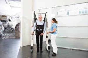 A man wearing a harness is supported by a robotic walking aid, while a woman in medical attire stands beside him in a clinical setting.