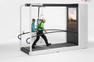 An elderly person uses a treadmill with support bars and a harness, assisted by a caregiver, with a scenic display screen in front of them.