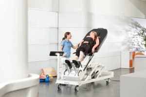 A healthcare professional assists a patient strapped to a tilt table in a bright clinical setting, likely for physical rehabilitation or therapy.