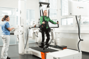 A person is strapped into a robotic gait therapy device while a clinician monitors the process on a control panel in a bright, modern rehabilitation room.