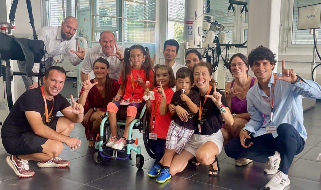 Un grupo de adultos y niños, algunos con distintivos con su nombre, posan en el interior y sonríen a la cámara; un niño utiliza una silla de ruedas.