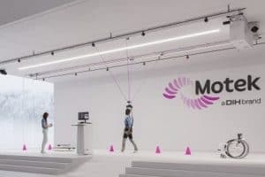 A person in a harness systems walks between pink cones in a clinical setting while another person monitors a computer; Motek logo is on the wall.