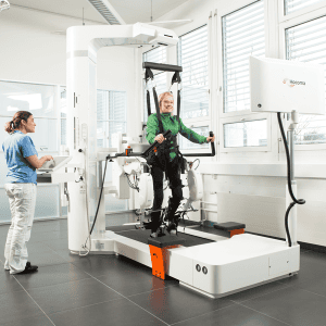 A woman in a green jacket uses a robotic gait trainer for rehabilitation, assisted by a healthcare professional in a clinical setting.