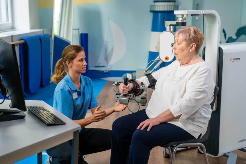 Un terapeuta ayuda a una mujer mayor a utilizar un dispositivo de brazo robótico para rehabilitación física en un entorno clínico.