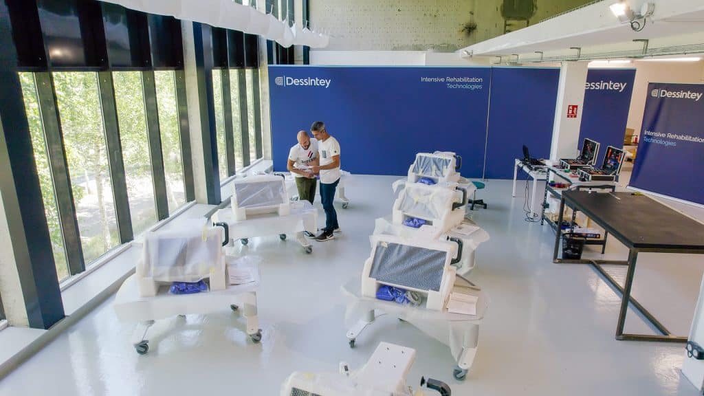 Dos personas se encuentran en una sala moderna y bien iluminada con varios dispositivos médicos cubiertos y estaciones de trabajo con el rótulo "Dessintey Intensive Rehabilitation Technologies".