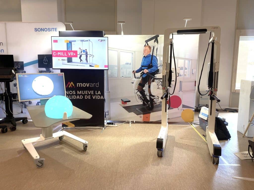 Se exponen equipos de rehabilitación médica, como una cinta de correr de realidad virtual, un monitor y un póster que muestra a una persona utilizando un dispositivo de ayuda a la movilidad.