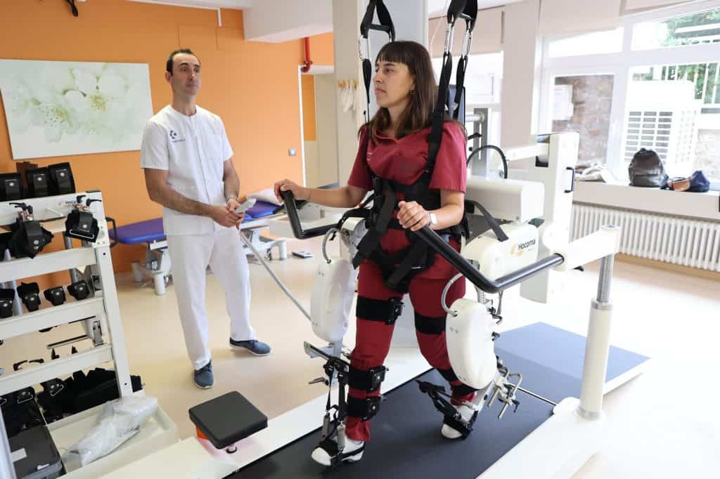 Una mujer utiliza un exoesqueleto robótico para rehabilitación física mientras un profesional sanitario observa en una sala de terapia.