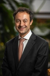 A man in a dark suit, white shirt, and light red tie is smiling and standing in front of a blurred outdoor background.