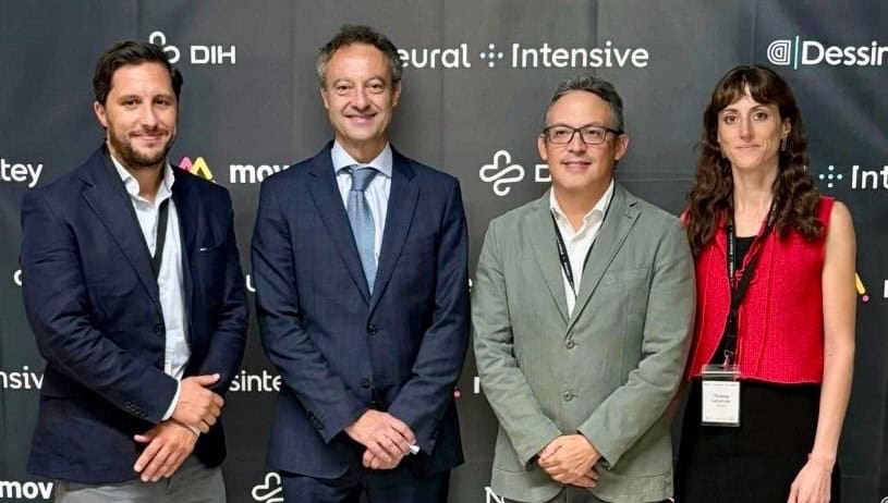 Cuatro personas de pie delante de un fondo negro con varios logotipos de empresas, vestidas con trajes de negocios y con insignias de conferencias.