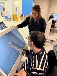 A man interacts with a virtual hand on a touchscreen while a woman stands beside him, providing guidance in a clinical or therapy setting.