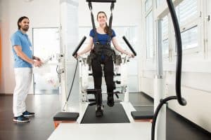 A woman uses a robotic exoskeleton treadmill for physical rehabilitation while a healthcare professional monitors the session.