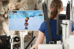 A person with shoulder straps uses rehabilitation equipment while playing a video game displayed on a monitor showing a snowy landscape and animated character.