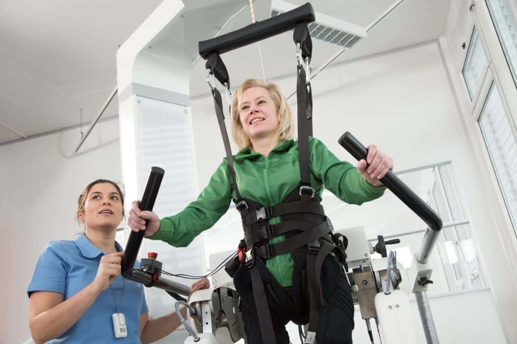 Una mujer con camisa verde utiliza una cinta de rehabilitación con soporte de arnés, asistida por un profesional sanitario con uniforme azul.