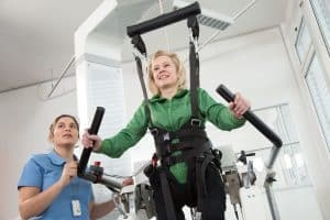 Una mujer con camisa verde utiliza una cinta de rehabilitación con soporte de arnés, asistida por un profesional sanitario con uniforme azul.