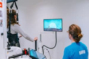 A person uses a rehabilitation machine while watching a screen, as another person in a blue shirt observes in a clinical setting.
