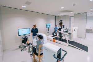 People use robotic rehabilitation equipment in a clinical setting; one person is on a treadmill device while another operates a seated arm machine, supervised by staff.