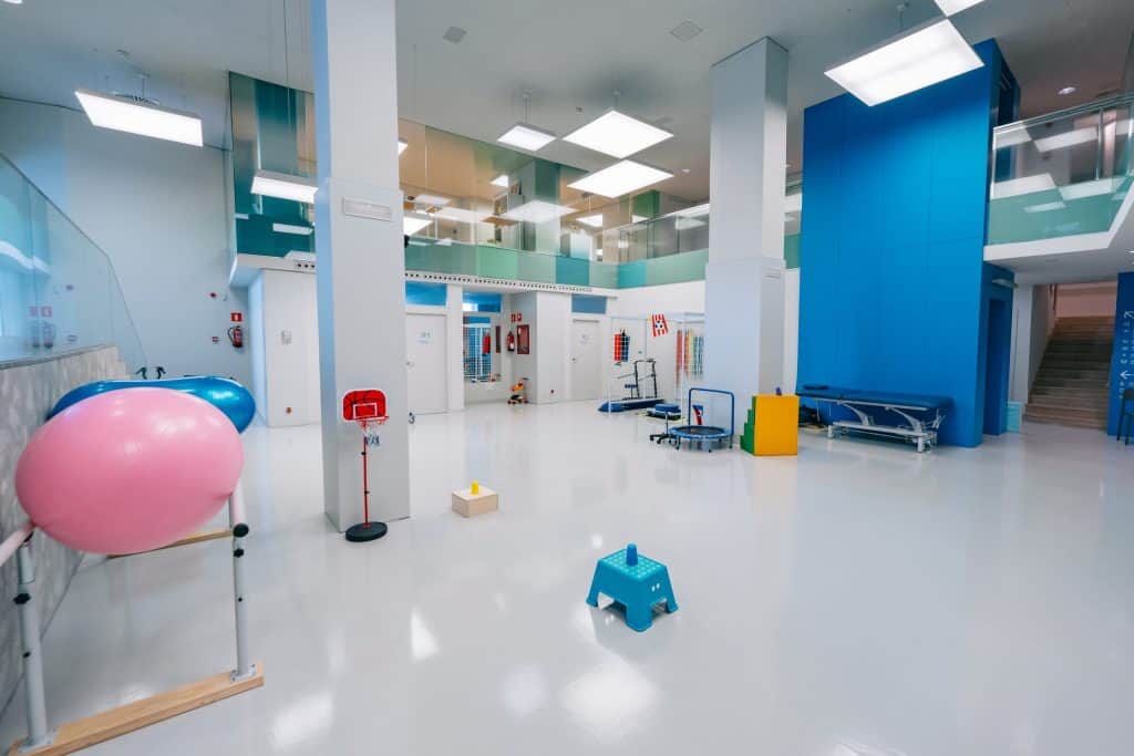 Una espaciosa sala de fisioterapia con pelotas de ejercicio, equipos terapéuticos, bancos y una iluminación brillante. Los suelos y las paredes son limpios y modernos, con colores azules y blancos por todas partes.