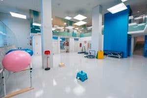 Una espaciosa sala de fisioterapia con pelotas de ejercicio, equipos terapéuticos, bancos y una iluminación brillante. Los suelos y las paredes son limpios y modernos, con colores azules y blancos por todas partes.